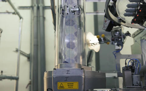 A close-up of a machine in a factory setting. The device features a tall, transparent cylinder containing several spheres. There are warning labels and wires nearby, with other mechanical components in the background.