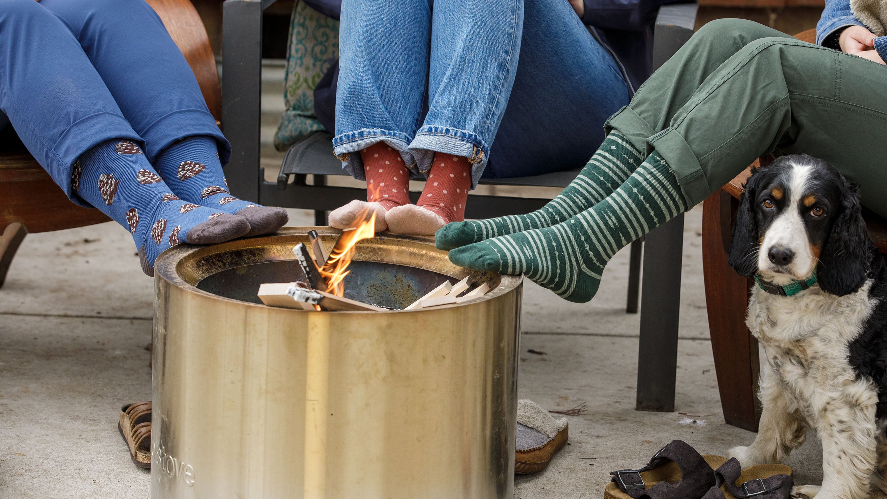 Three people with colorful socks relax by a small fire pit, with a black and white dog sitting nearby. The scene conveys a cozy atmosphere outdoors.